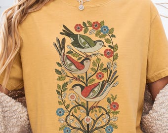 May include: Mustard yellow t-shirt featuring a detailed illustration of three birds perched on a flowering branch. The birds have red, green, and beige coloring, with floral accents in red, blue, and white. The shirt is a comfortable, casual style.