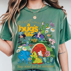 May include: Green t-shirt featuring a colorful graphic from "A Bug's Life." The design includes various insect characters, flowers, and the text "World Famous a bug's life" and "Bug-sized fun for everyone!"