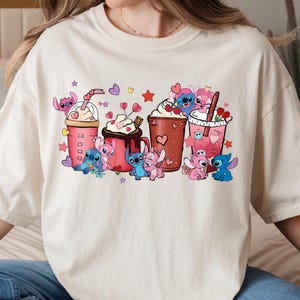 May include: Cream-colored t-shirt featuring a colorful graphic of cartoon characters and various beverages. The design includes multiple cups and mugs with whipped cream, hearts, and stars. The cartoon characters are in various poses, adding a playful touch to the design.