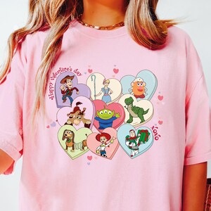 May include: Pink t-shirt featuring a colorful graphic of Toy Story characters inside heart shapes. The phrase "Happy Valentine's Day" is printed on the shirt, along with "xoxo". The design includes Woody, Buzz Lightyear, and other characters.