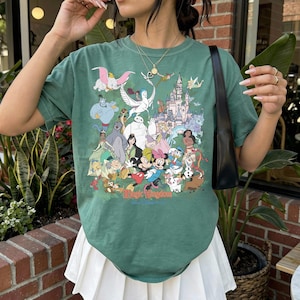 May include: A sage green short-sleeve t-shirt featuring a colorful graphic print of various animated characters and a castle. The text "Magic Kingdom" is printed in red at the bottom of the graphic. The shirt has a crew neckline and a relaxed fit.