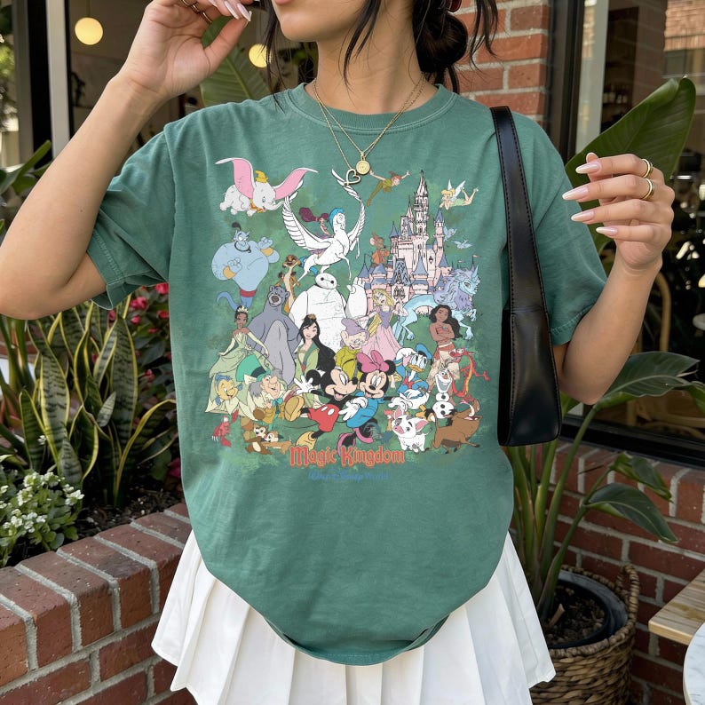 May include: A sage green short-sleeve t-shirt featuring a colorful graphic print of various animated characters and a castle. The text "Magic Kingdom" is printed in red at the bottom of the graphic. The shirt has a crew neckline and a relaxed fit.