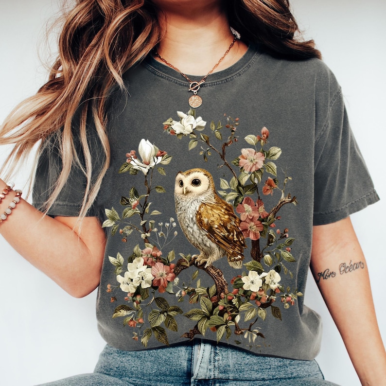 K&ouml;nnte beinhalten: Dunkelgraues T-Shirt mit einer detaillierten Illustration einer Eule auf einem bl&uuml;henden Ast. Das Design umfasst wei&szlig;e, rosa und gr&uuml;ne florale Elemente. Das T-Shirt hat einen Vintage-Look.