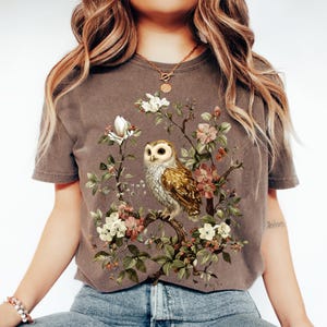 K&ouml;nnte beinhalten: Braunes T-Shirt mit detaillierter Illustration einer Eule auf einem bl&uuml;henden Ast. Das Design umfasst wei&szlig;e, rosa und gr&uuml;ne florale Elemente. Das T-Shirt hat ein Vintage-Aussehen, leicht verblichen.