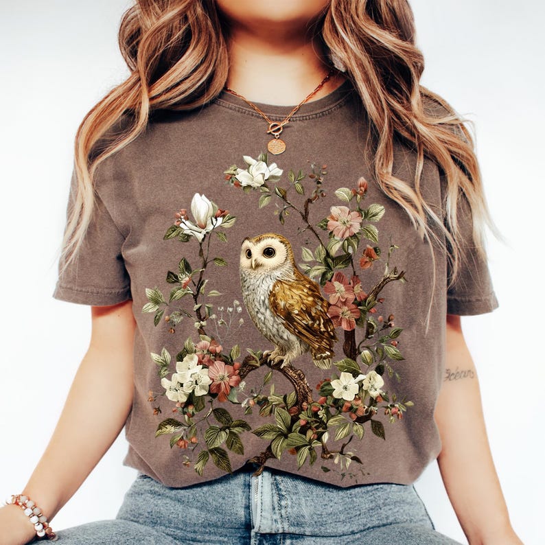K&ouml;nnte beinhalten: Braunes T-Shirt mit detaillierter Illustration einer Eule auf einem bl&uuml;henden Ast. Das Design umfasst wei&szlig;e, rosa und gr&uuml;ne florale Elemente. Das T-Shirt hat ein Vintage-Aussehen, leicht verblichen.