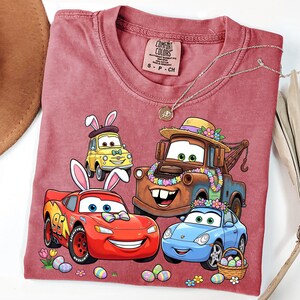 May include: A maroon Comfort Colors t-shirt featuring a graphic of animated cars with bunny ears and Easter eggs. The cars include a yellow Fiat, a red race car, a brown tow truck, and a blue Porsche. A silver necklace with a pendant is also visible.