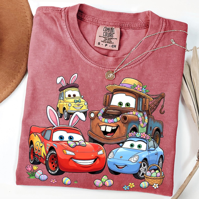 May include: A maroon Comfort Colors t-shirt featuring a graphic of animated cars with bunny ears and Easter eggs. The cars include a yellow Fiat, a red race car, a brown tow truck, and a blue Porsche. A silver necklace with a pendant is also visible.