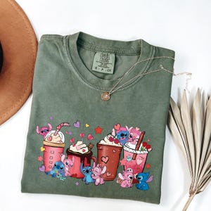 May include: Olive green t-shirt with a graphic of cartoon characters and beverages. The design includes pink and blue characters with hearts and stars, alongside pink and brown drink cups. A gold necklace with a pendant is draped across the shirt.