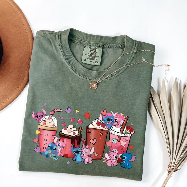 May include: Olive green t-shirt with a graphic of cartoon characters and beverages. The design includes pink and blue characters with hearts and stars, alongside pink and brown drink cups. A gold necklace with a pendant is draped across the shirt.