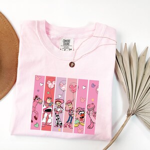 Op de afbeelding: Een lichtroze Comfort Colors T-shirt met een afbeelding van tekenfilmfiguren uit een populaire animatiefilm. De afbeelding is verdeeld in verticale roze strepen, met harten en Mickey Mouse-ballonnen. Een gouden ketting met een hanger is over het shirt gedrapeerd.