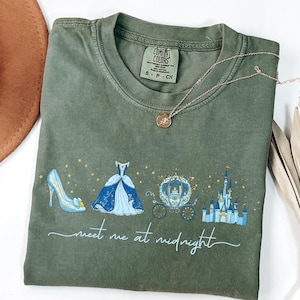 May include: Olive green t-shirt featuring a Cinderella-themed design with a glass slipper, ball gown, carriage, and castle. The text "meet me at midnight" is printed below the illustrations. A gold necklace with a pendant is draped on the shirt.