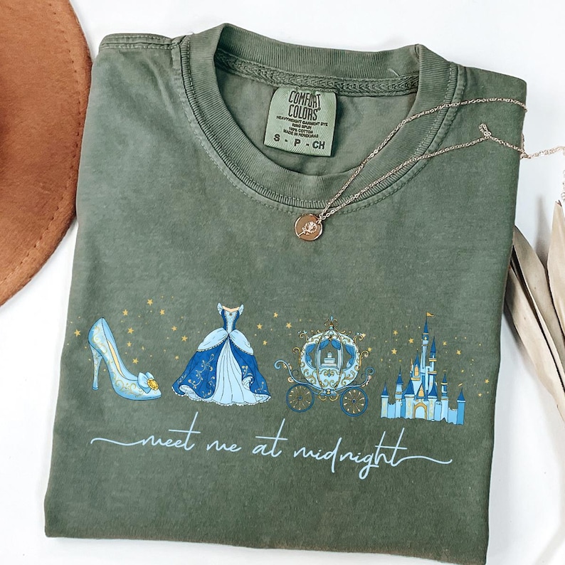 May include: Olive green t-shirt featuring a Cinderella-themed design with a glass slipper, ball gown, carriage, and castle. The text "meet me at midnight" is printed below the illustrations. A gold necklace with a pendant is draped on the shirt.