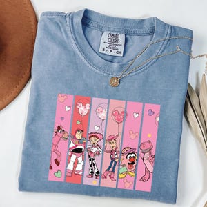 Op de afbeelding: Een lichtblauw Comfort Colors t-shirt met een afbeelding van personages uit een populaire animatiefilm, waaronder Woody, Buzz Lightyear en Jessie. Het ontwerp heeft roze verticale strepen en Mickey Mouse ballonaccenten. Een gouden ketting ligt over het gevouwen t-shirt.