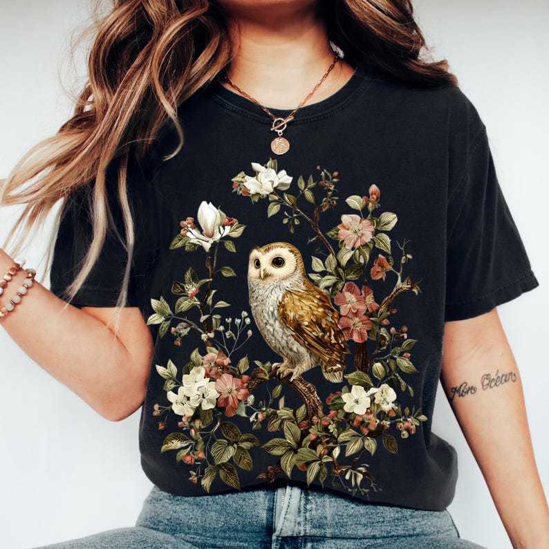 K&ouml;nnte beinhalten: Schwarzes T-Shirt mit einer detaillierten Illustration einer Eule, die auf einem bl&uuml;henden Ast sitzt. Das Design umfasst wei&szlig;e, rosa und gr&uuml;ne florale Elemente, die eine Vintage-&Auml;sthetik erzeugen. Die Eule hat braune und wei&szlig;e Federn.