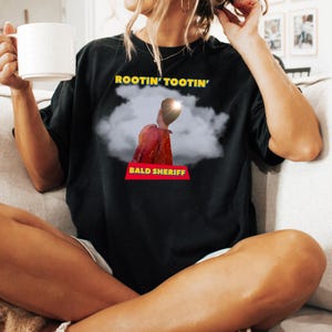 May include: Black t-shirt featuring the text "ROOTIN' TOOTIN'" in yellow above a graphic of a person in a red jacket and the text "BALD SHERIFF" in a red banner. The person is set against a cloud background.