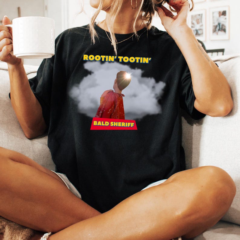 May include: Black t-shirt featuring the text "ROOTIN' TOOTIN'" in yellow above a graphic of a person in a red jacket and the text "BALD SHERIFF" in a red banner. The person is set against a cloud background.
