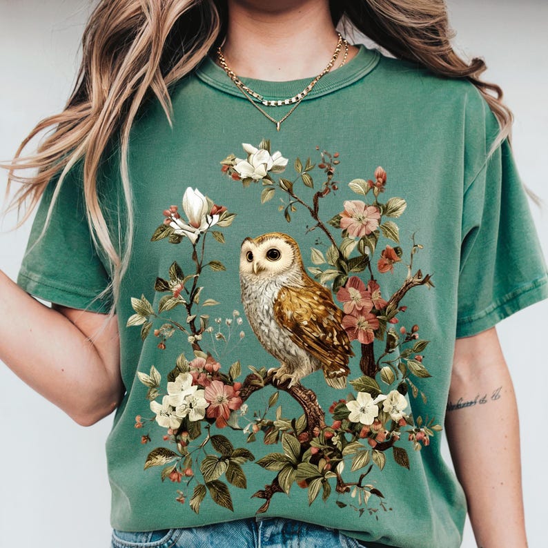K&ouml;nnte beinhalten: Gr&uuml;nes T-Shirt mit einer detaillierten Illustration einer Eule, die auf einem bl&uuml;henden Ast sitzt. Die Eule hat braune und wei&szlig;e Federn, umgeben von rosa, wei&szlig;en und cremefarbenen Bl&uuml;ten. Das Shirt hat eine einfarbige gr&uuml;ne Farbe.