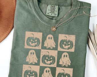 May include: Olive green t-shirt with a grid pattern of tan pumpkins and ghosts. The shirt has a crew neck and short sleeves. A silver necklace with a small pendant is draped across the shirt. The tag reads "Comfort Colors".