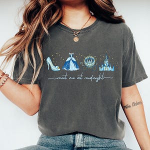 May include: Dark gray t-shirt featuring a Cinderella-themed design. The graphic includes a glass slipper, ball gown, carriage, and castle, with the text "meet me at midnight" in a script font. The design is in shades of blue and gold.