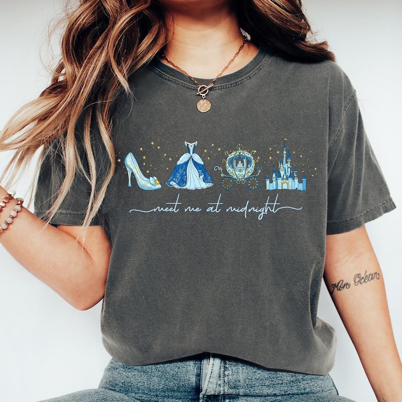 May include: Dark gray t-shirt featuring a Cinderella-themed design. The graphic includes a glass slipper, ball gown, carriage, and castle, with the text "meet me at midnight" in a script font. The design is in shades of blue and gold.