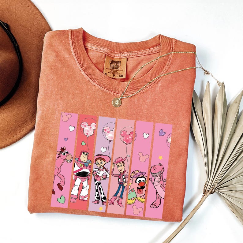 Op de afbeelding: Koraalrood T-shirt met een afbeelding van personages uit een populaire animatiefilm. Het ontwerp bevat een roze en witte gestreepte achtergrond met hart- en Mickey Mouse ballonaccenten. Een gouden ketting en een bruine hoed zijn ook zichtbaar.