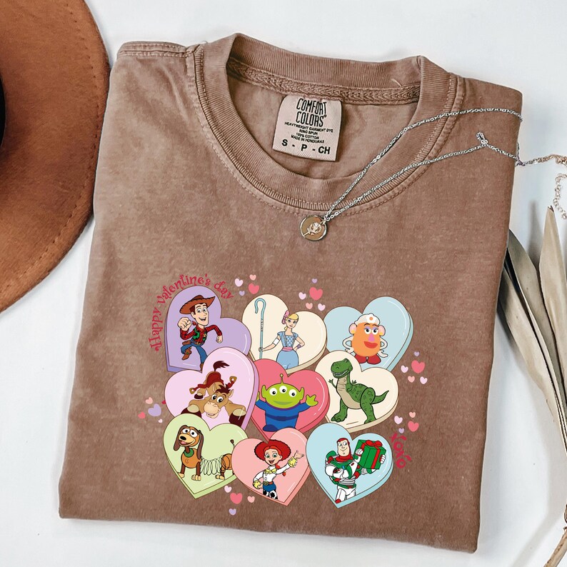 May include: Brown t-shirt with a graphic of Toy Story characters in heart shapes. The phrase "Happy Valentine's Day" is printed on the shirt. A silver necklace with a small pendant is draped across the shirt.