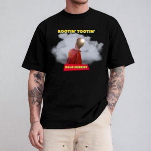 May include: Black t-shirt featuring the text "ROOTIN' TOOTIN'" in yellow above an image of a person in a red jacket and a cloud. Below the image is the text "BALD SHERIFF" in a red rectangle.