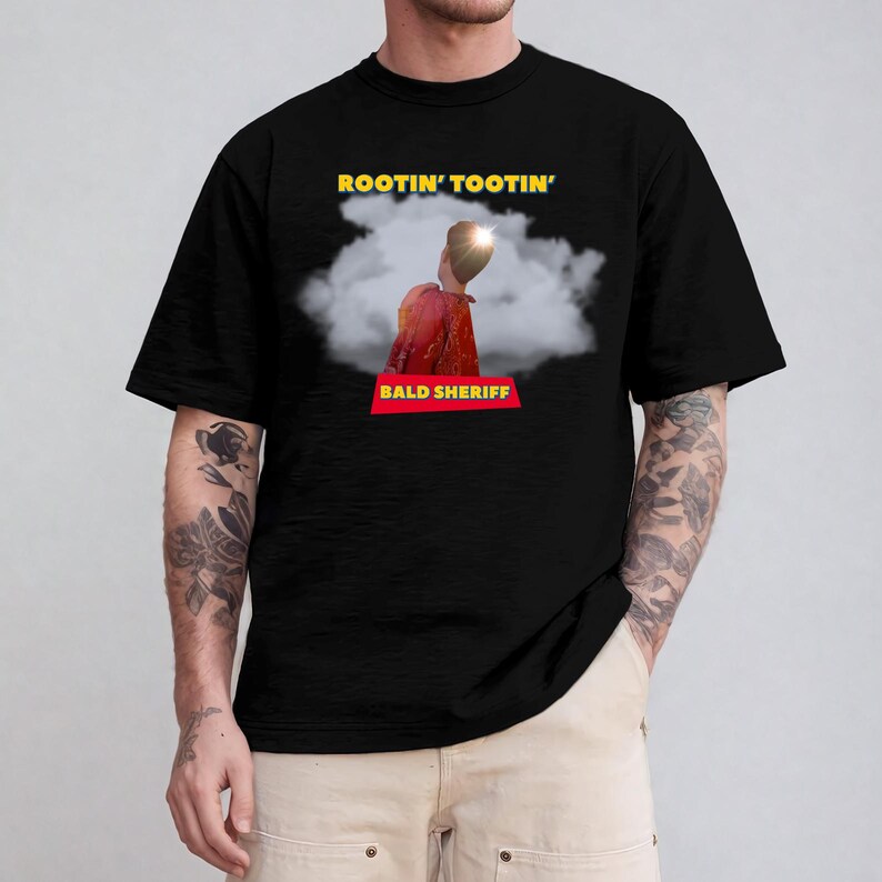May include: Black t-shirt featuring the text "ROOTIN' TOOTIN'" in yellow above an image of a person in a red jacket and a cloud. Below the image is the text "BALD SHERIFF" in a red rectangle.