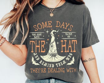 May include: Dark gray t-shirt with orange and cream-colored text and graphics. The text reads "SOME DAYS YOU JUST HAVE TO PUT ON THE HAT AND REMIND THEM WHO THEY'RE DEALING WITH." A witch hat and spiderweb graphic is in the center.