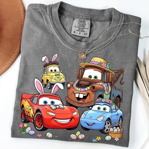 May include: A gray Comfort Colors t-shirt featuring a colorful graphic of animated cars with Easter bunny ears and floral decorations. The cars include a red race car, a tow truck, a yellow car, and a blue car. The shirt also has a small tag.