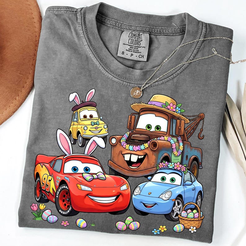 May include: A gray Comfort Colors t-shirt featuring a colorful graphic of animated cars with Easter bunny ears and floral decorations. The cars include a red race car, a tow truck, a yellow car, and a blue car. The shirt also has a small tag.