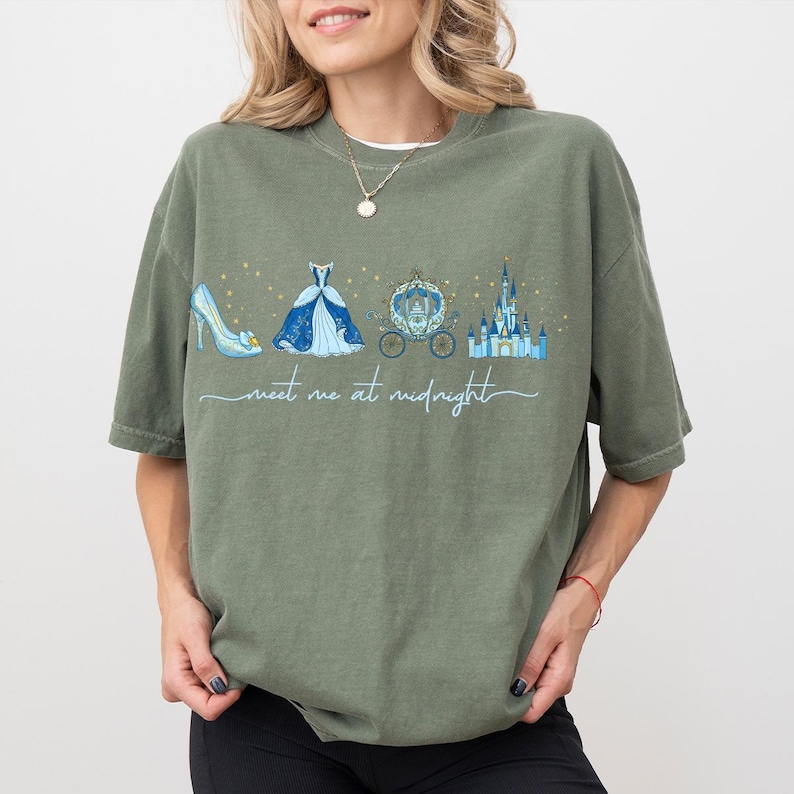 May include: Olive green t-shirt featuring a Cinderella-themed graphic. The design includes a blue glass slipper, a ball gown, a carriage, and a castle, with the text "meet me at midnight" in a script font.