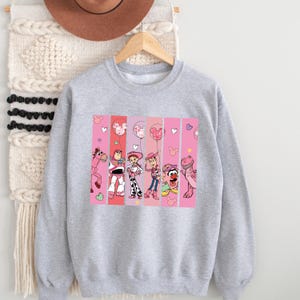Op de afbeelding: Een lichtgrijze sweatshirt met een kleurrijke afbeelding van stripfiguren. De afbeelding is verdeeld in verticale roze panelen met afbeeldingen van een paard, Buzz Lightyear, Jessie, Woody en andere personages. Er zijn ook harten en ballonnen.