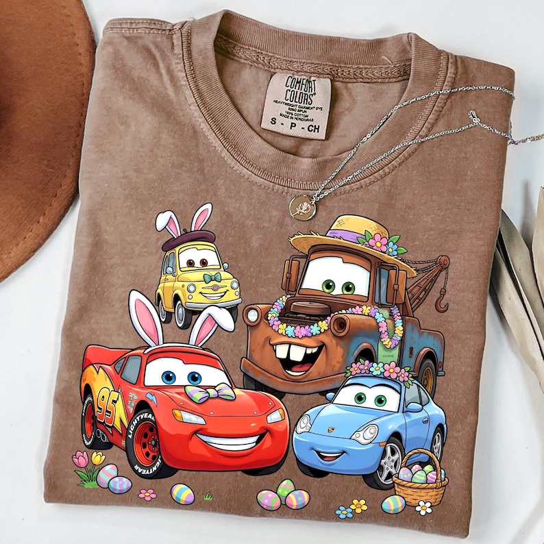 May include: Brown t-shirt featuring cartoon cars with bunny ears and Easter eggs. The shirt has the text "Comfort Colors" on the tag. A silver necklace is draped across the shirt.