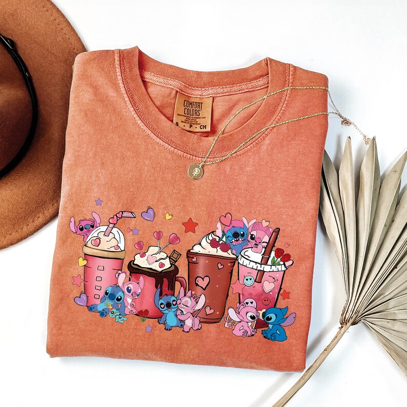 May include: Coral-colored Comfort Colors t-shirt featuring a graphic of Stitch characters with various drinks, hearts, and stars. A gold necklace and a brown hat are visible. The shirt has a crew neck and short sleeves.