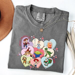 May include: A gray Comfort Colors t-shirt featuring a graphic of Toy Story characters inside heart shapes. The words "Happy Valentine's Day" are printed on the shirt. A silver necklace with a small pendant is draped across the shirt.