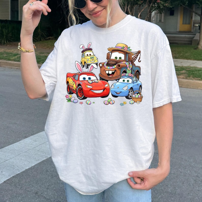 May include: White t-shirt featuring a colorful graphic of animated cars decorated for Easter. The design includes a red car with bunny ears, a brown tow truck, a yellow car, and a blue car, surrounded by Easter eggs and flowers.