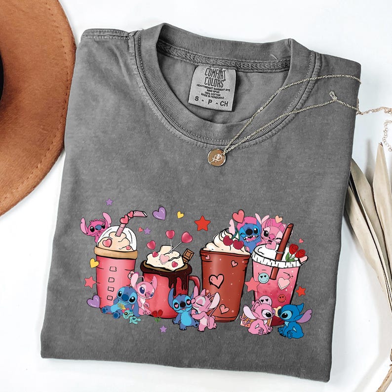 May include: A gray Comfort Colors t-shirt featuring a colorful graphic of cartoon characters with coffee drinks and hearts. The shirt has a crew neck and short sleeves. A silver necklace with a small pendant is on the shirt.