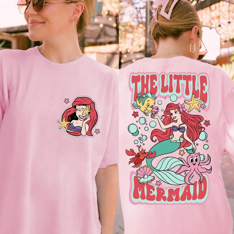 Może przedstawiać: Jasnor&oacute;żowa koszulka z kresk&oacute;wkową ilustracją syreny. Z przodu mały obrazek syreny, a z tyłu większa ilustracja z napisem "THE LITTLE MERMAID" i stworzeniami morskimi.
