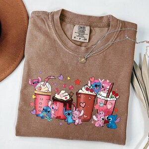May include: Brown t-shirt with a graphic of cartoon characters and drinks. The graphic includes pink, blue, and white characters with various drinks, hearts, and stars. The shirt has a crew neck and a small tag.