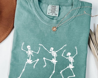 May include: A teal Comfort Colors t-shirt featuring a graphic of three dancing skeletons. The shirt has a crew neck and short sleeves. A silver necklace with a small pendant rests on the shirt. The shirt is folded on a white surface.
