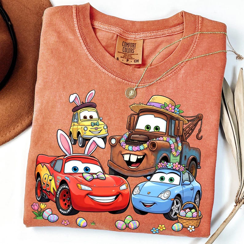 May include: Orange Comfort Colors t-shirt featuring cartoon cars decorated for Easter. The design includes Lightning McQueen, Mater, Sally, and a yellow car, all with Easter bunny ears and floral decorations. A gold necklace is draped across the shirt.