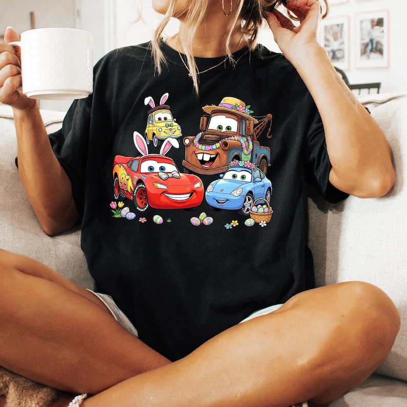May include: Black t-shirt featuring a colorful graphic of animated cars decorated for Easter. The design includes a red race car, a blue car, a yellow car, and a brown tow truck, all with Easter-themed accessories and surrounded by Easter eggs.