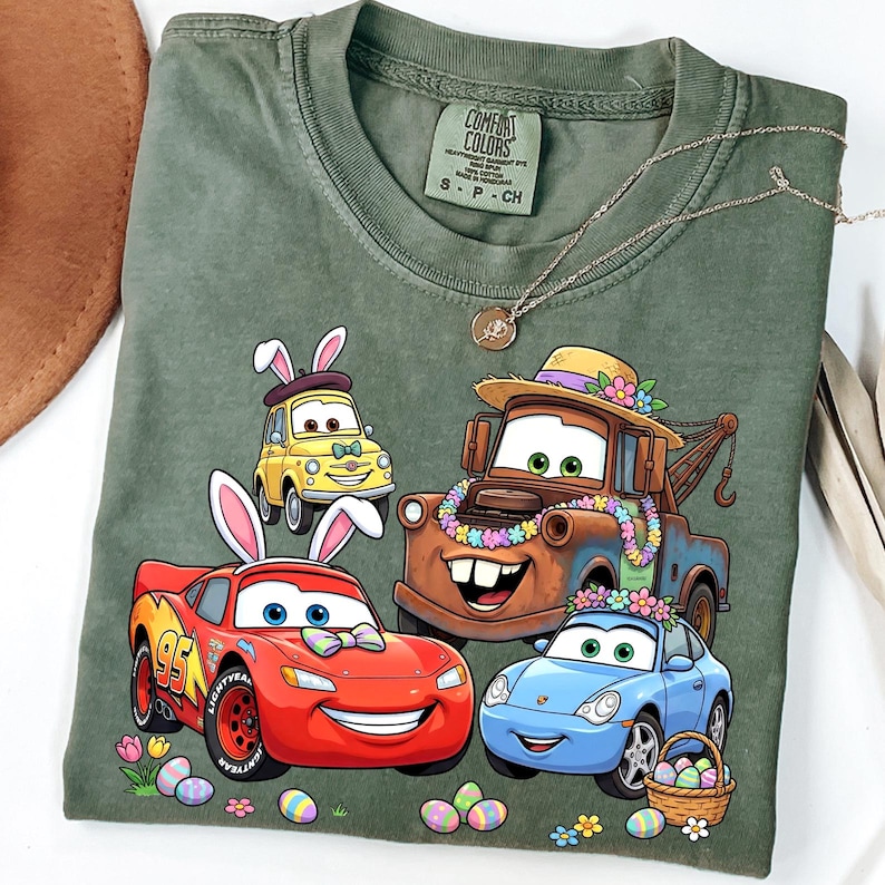 May include: Green t-shirt featuring cartoon cars with bunny ears and Easter eggs. The design includes a red car with the number 95, a yellow car, a brown tow truck, and a blue car. The shirt has a Comfort Colors tag.