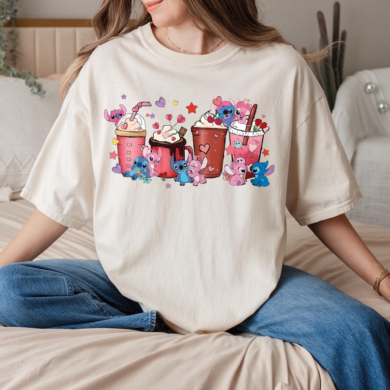 May include: Cream-colored t-shirt featuring a colorful graphic of cartoon characters and various beverages. The design includes multiple cups and mugs with whipped cream, hearts, and stars. The cartoon characters are in various poses, adding a playful touch to the design.