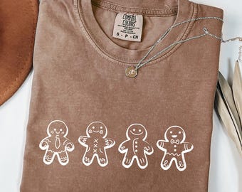May include: A brown Comfort Colors t-shirt featuring a white graphic of four gingerbread men. The shirt has a crew neck and short sleeves. A silver necklace with a small pendant is draped across the shirt.
