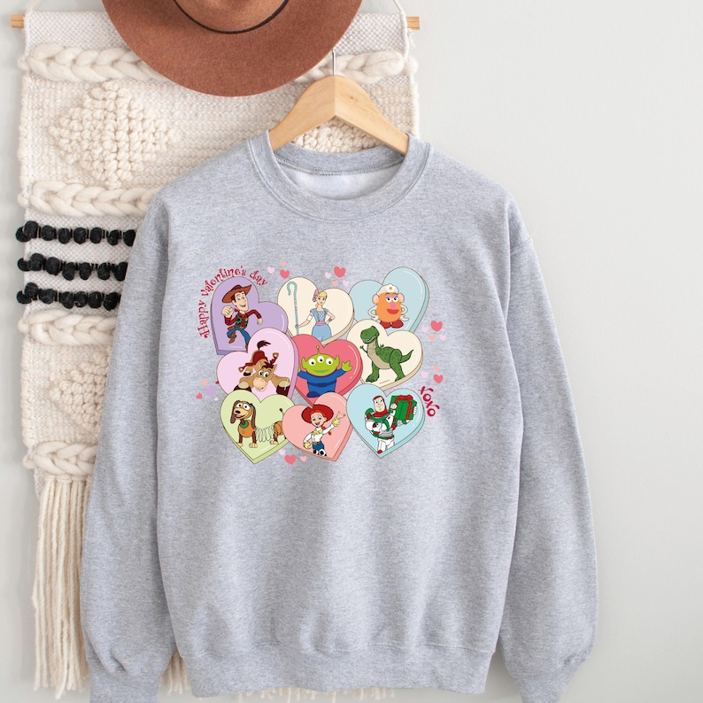 May include: A light gray sweatshirt featuring a colorful graphic of Toy Story characters inside heart shapes. The words "Happy Valentine's Day" are printed on the left side, with "xoxo" on the right. The sweatshirt is hanging on a wooden hanger.
