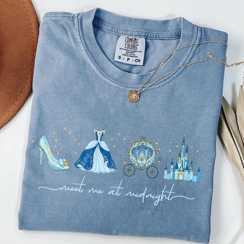 May include: A blue Comfort Colors t-shirt featuring a Cinderella-themed design. The design includes a high-heeled shoe, a ball gown, a carriage, and a castle, with the words "meet me at midnight" in script. A gold necklace rests on the shirt.