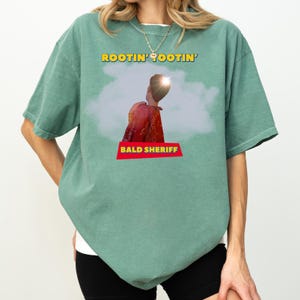 May include: Sage green t-shirt featuring the text "ROOTIN' TOOTIN'" in yellow and a graphic of a person in a red shirt. The text "BALD SHERIFF" is in a red banner below the graphic. The shirt has short sleeves and a crew neck.