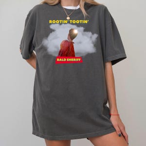 May include: Charcoal grey t-shirt featuring the text "ROOTIN' TOOTIN'" in yellow above an image of a person in a red shirt. The text "BALD SHERIFF" is in a red box below the image.